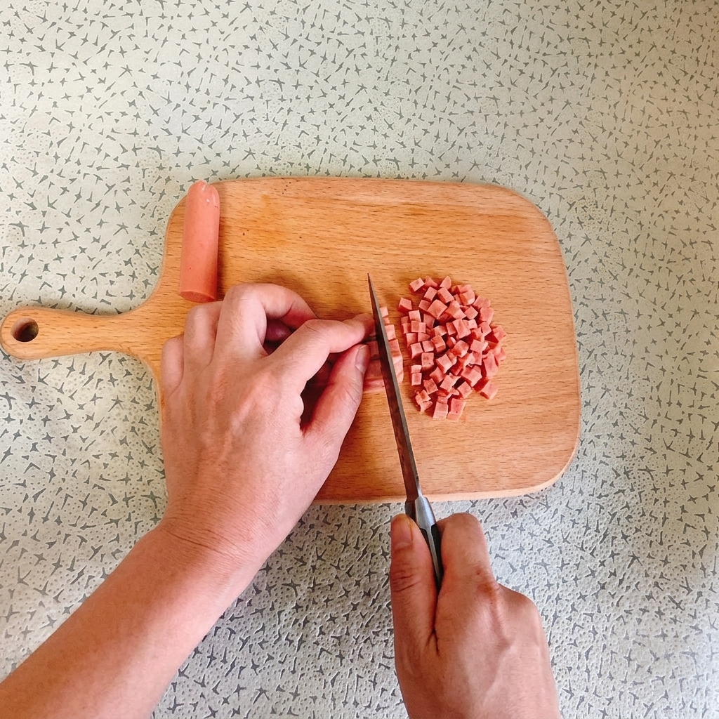 A persons hands using a chefs knife to dice a pink ham sausage into small, even cubes on a light wooden cutting board.