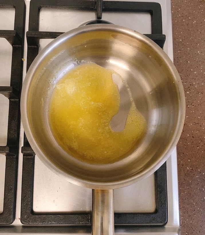 A stainless steel saucepan on a gas stove containing melted yellow butter with a spoonful of white flour being added to the center.