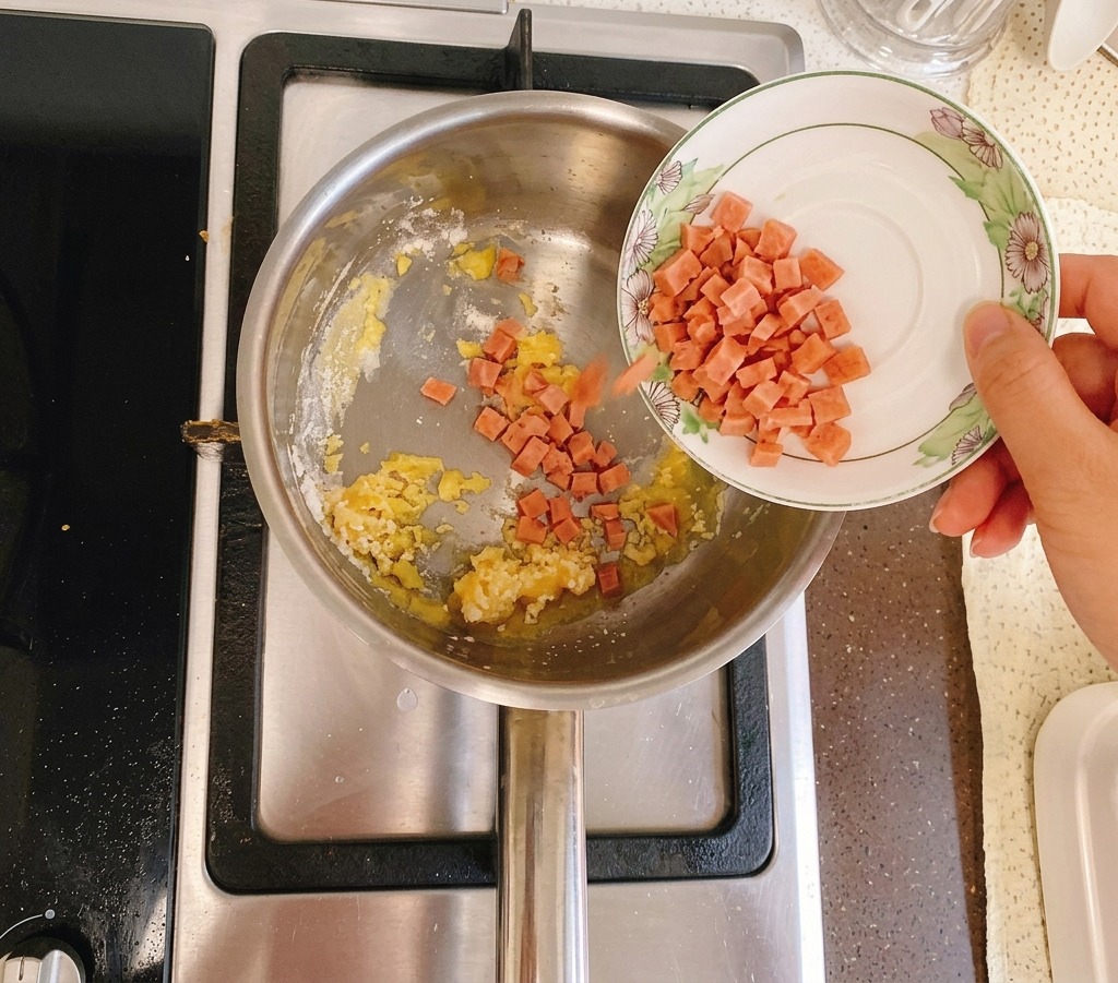 Diced ham being poured from a small white plate into a pot containing a golden butter and flour mixture.