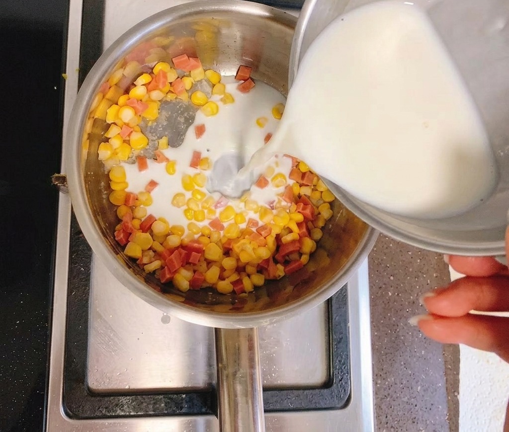 A person pouring cloudy corn water from a silver bowl into a saucepan containing yellow corn kernels and diced ham.