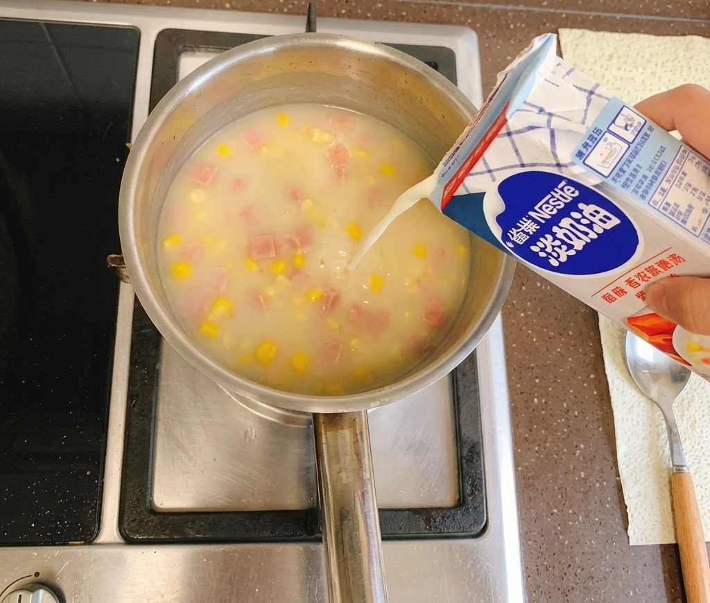 Light cream being poured from a blue and white carton into a simmering pot of creamy corn soup on a stovetop.