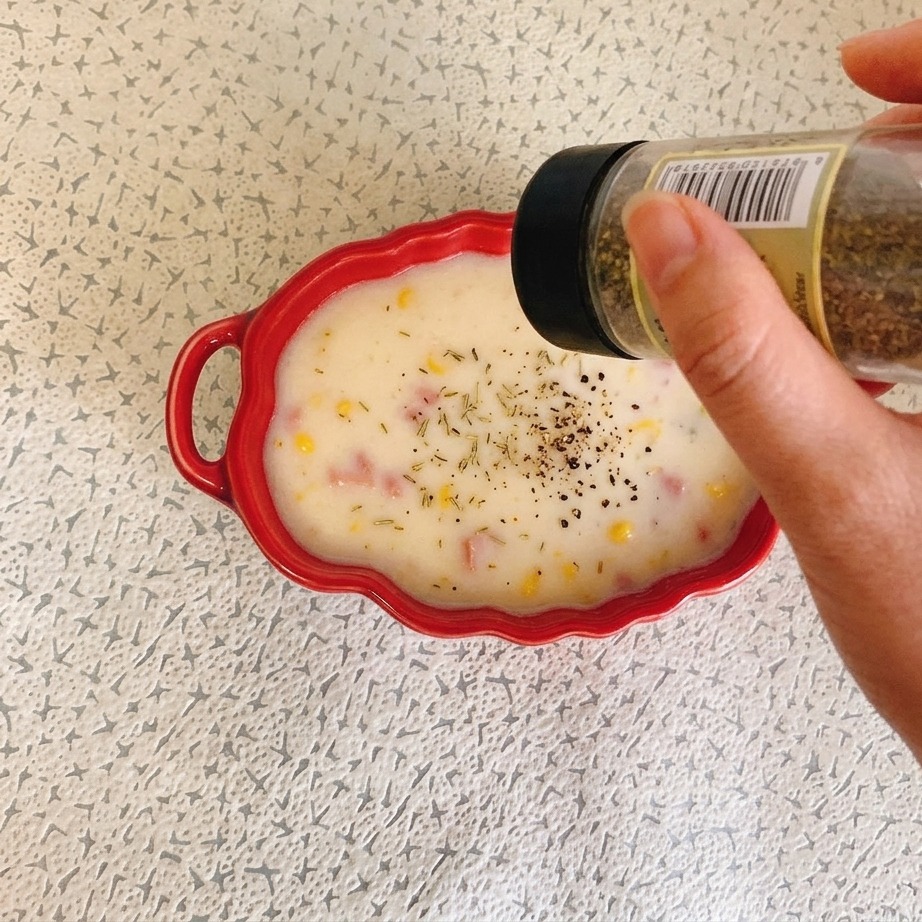 A hand holding a spice jar and sprinkling dried herbs onto a red bowl of creamy ham and corn soup.