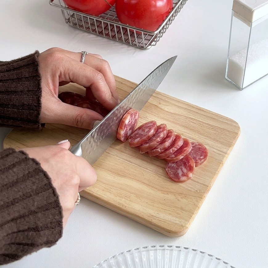 Hands using a silver kitchen knife to slice a red sausage into thin circular rounds on a light-colored wooden cutting board.