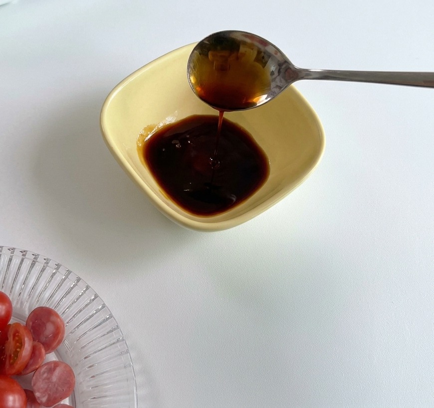 A metal spoon pouring dark soy sauce into a yellow prep bowl that already contains a portion of red tomato sauce.