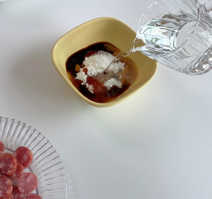 Clear water being poured from a glass pitcher into a yellow bowl containing various sauces and a mound of white cornstarch.