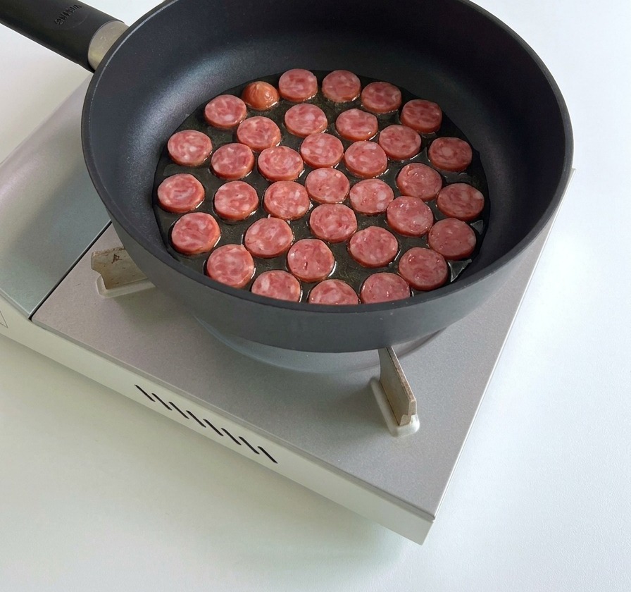 Numerous sliced sausage rounds being fried in a single layer in a black pan on a portable stove.