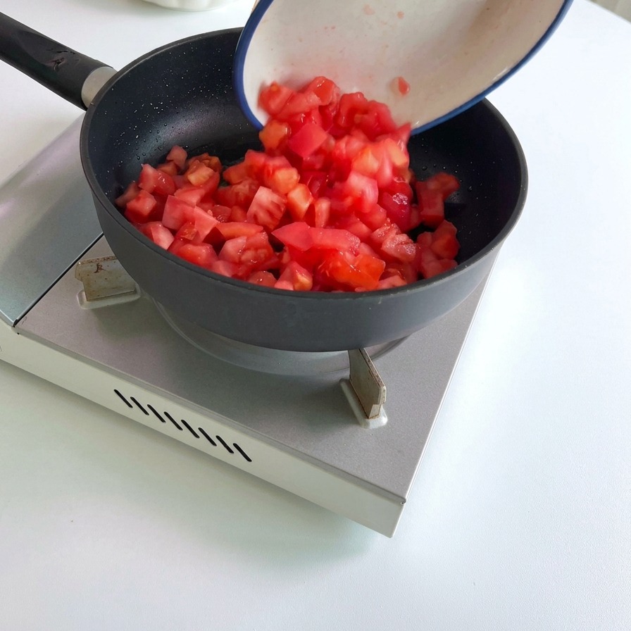 Diced red tomatoes being poured from a white ceramic bowl into a hot black frying pan.