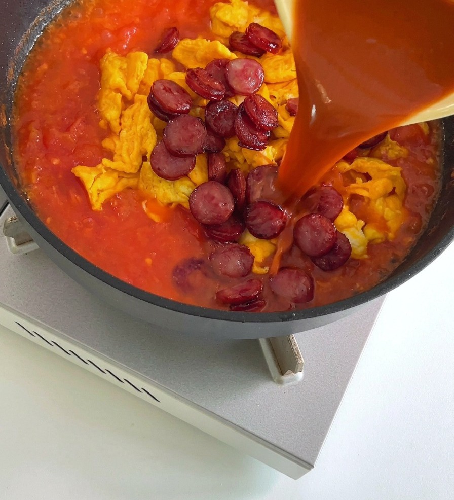 A reddish sauce being poured from a yellow bowl into a frying pan containing scrambled eggs and sliced sausages in a tomato-based mixture.
