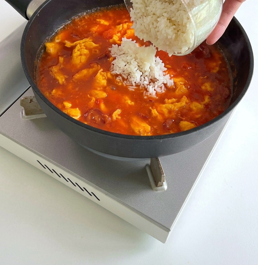 White cooked rice being tipped from a glass bowl into a pan of bubbling tomato sauce, eggs, and sausage.