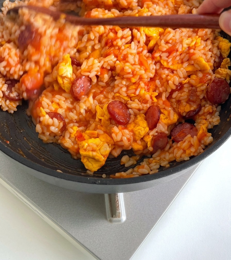 Rice being mixed with a wooden cooking utensil in a frying pan with tomato sauce, sausages, and eggs until fully integrated.