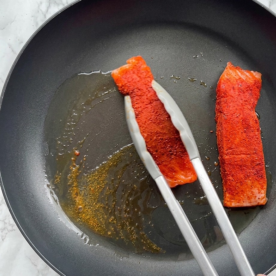 A seasoned salmon fillet being seared in a hot pan with bubbling olive oil, held by metal tongs.