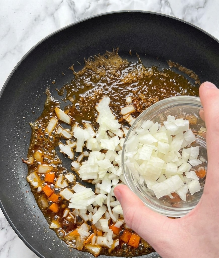 Diced white onions being poured from a glass bowl into a pan containing oil and salmon pan-drippings.