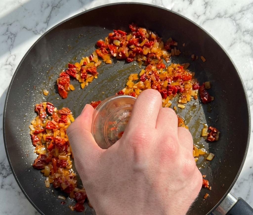 A hand pouring chopped sun-dried tomatoes and red pepper flakes from a small glass bowl into a pan of sautéing aromatics.