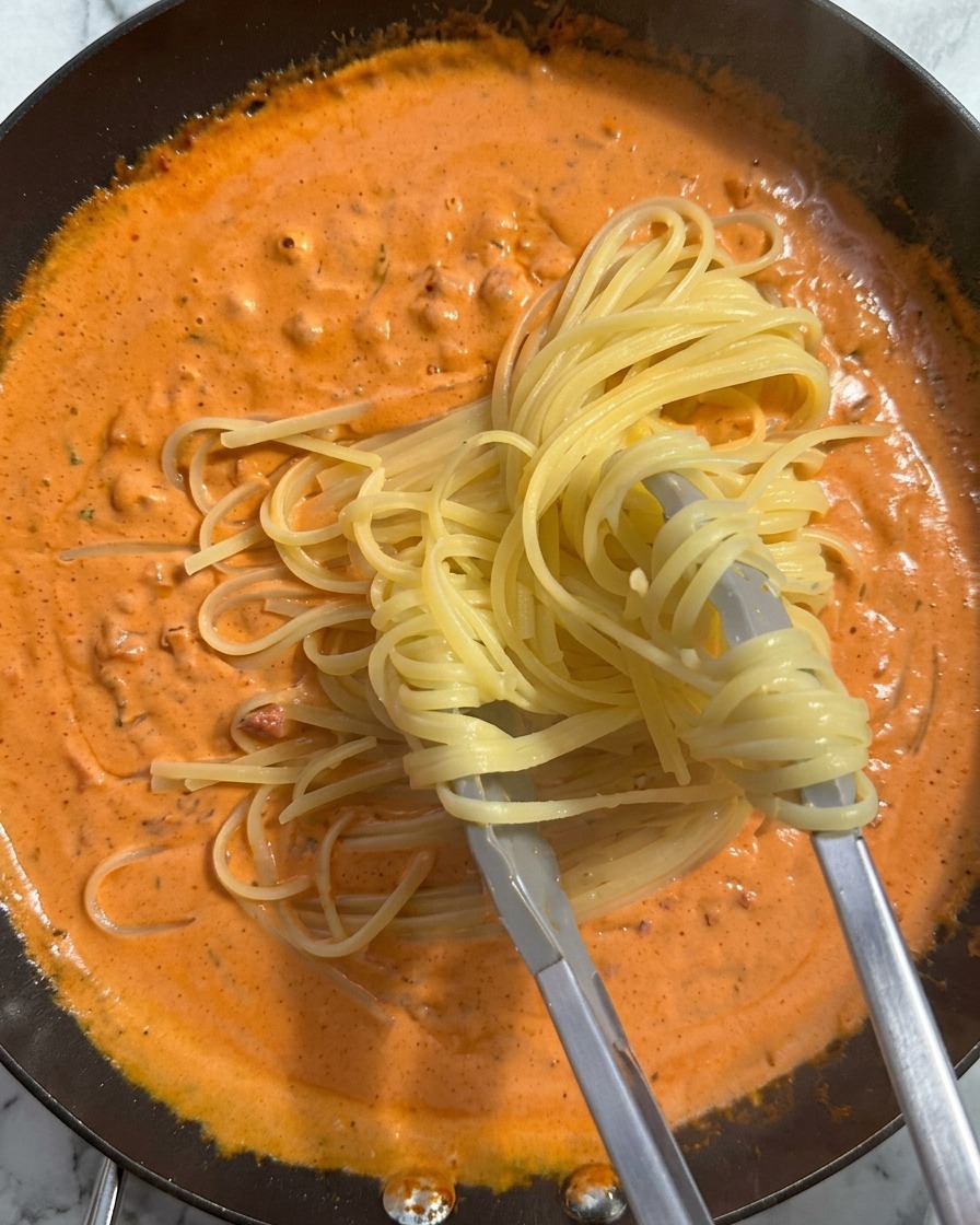 A bundle of cooked pasta being lifted by tongs and placed into a pan filled with a rich, creamy orange sauce.