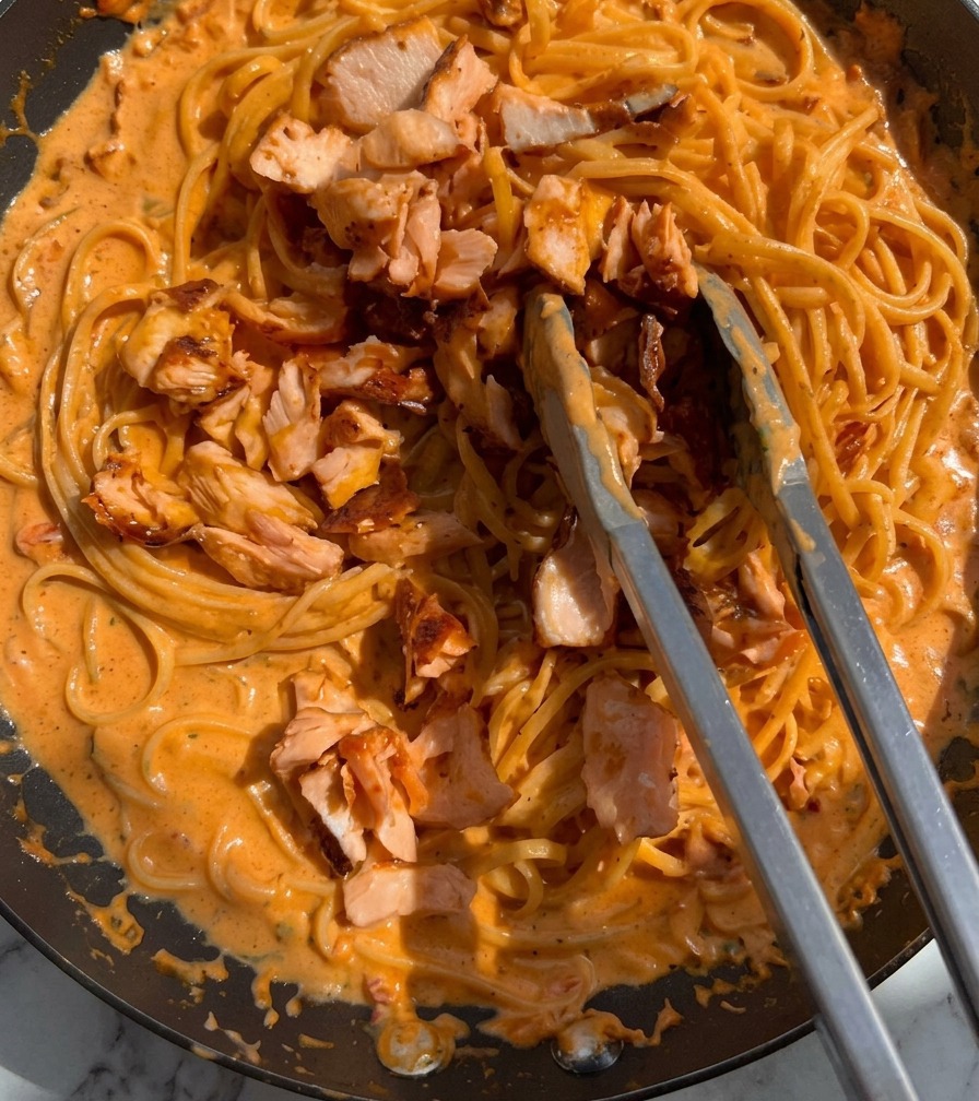 Pieces of cooked pink salmon being tossed into a pan of creamy pasta using metal tongs.
