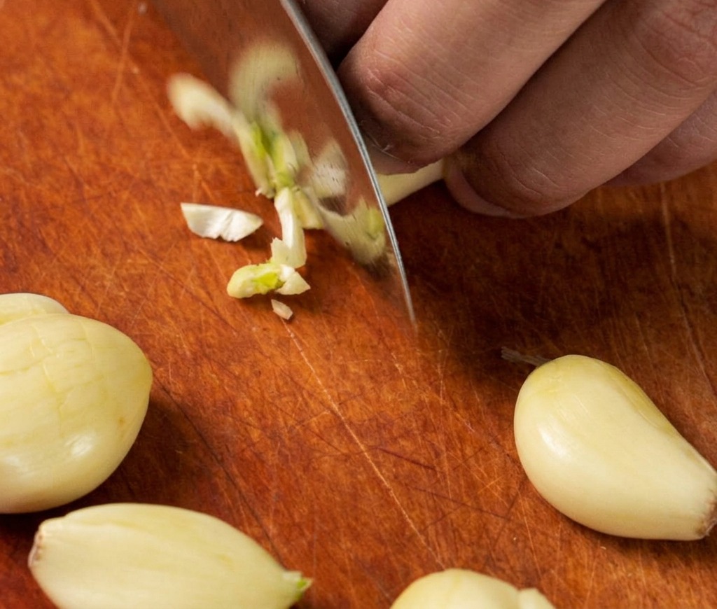 A close-up view of a chefs knife mincing several cloves of fresh garlic into small pieces on a wooden cutting board.