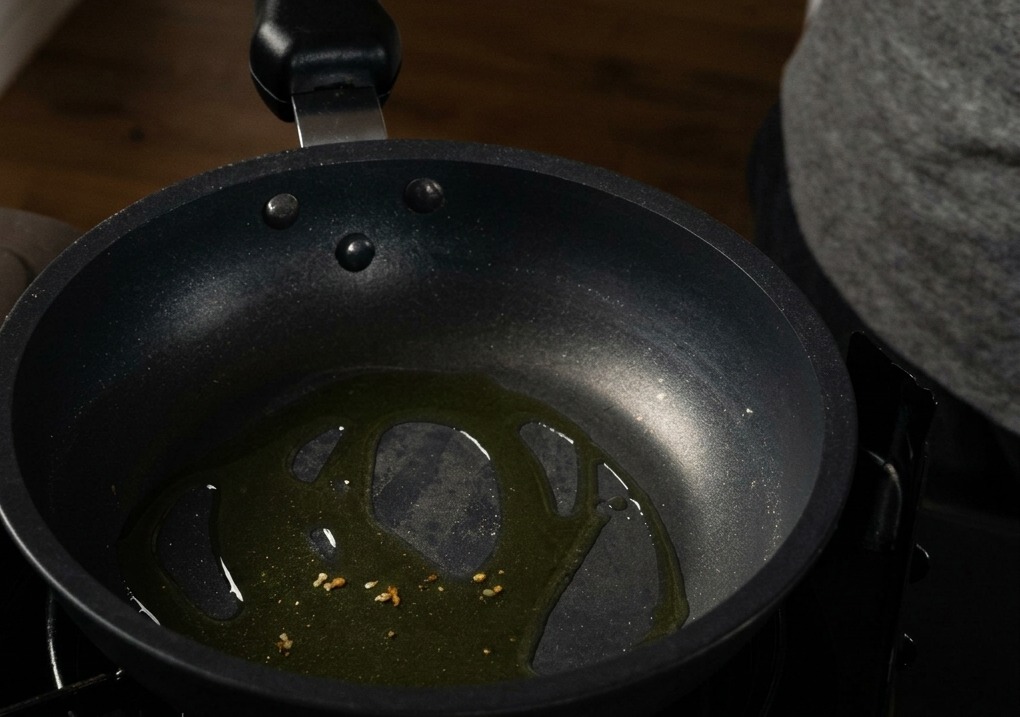 Minced garlic pieces sizzling in olive oil at the bottom of a black non-stick frying pan.