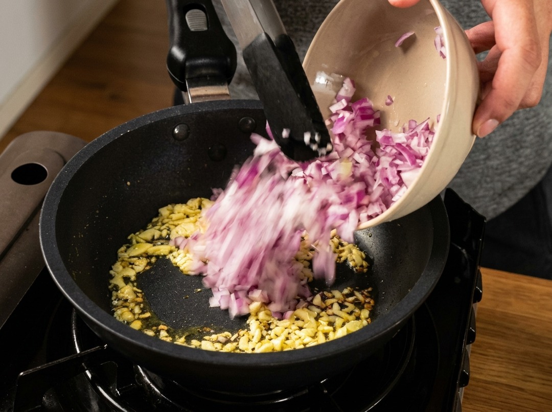 Diced purple onions being poured from a bowl into a frying pan to be sautéed with garlic.
