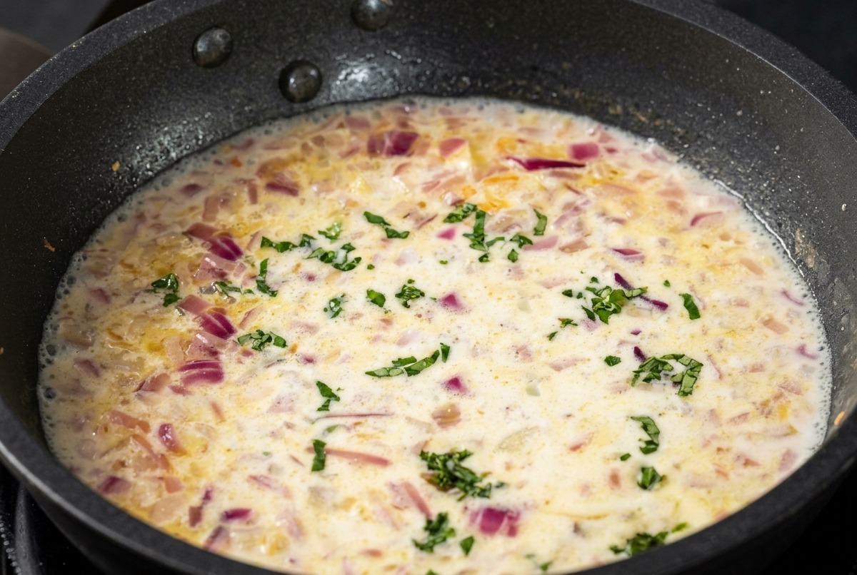 A creamy white sauce in a skillet with flecks of green minced basil scattered throughout the mixture.