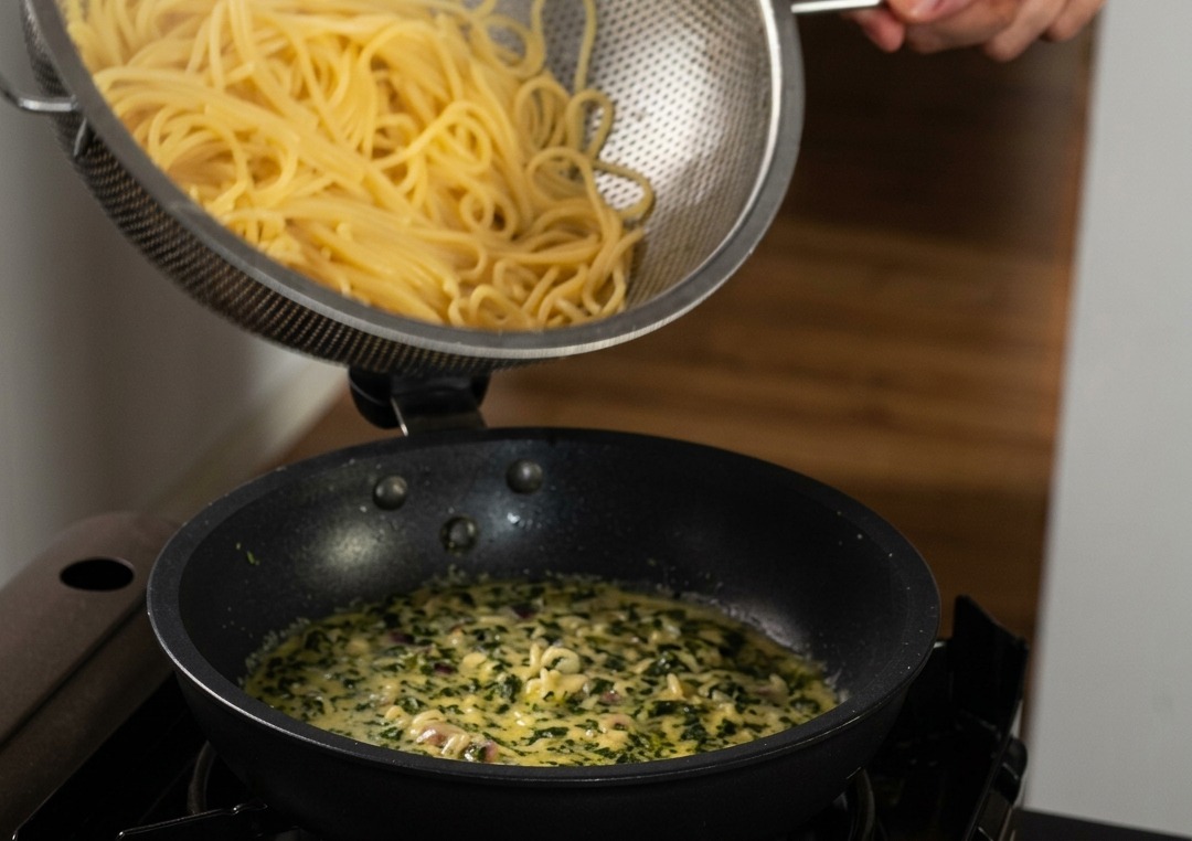 Cooked spaghetti being poured from a stainless steel strainer into a pan of creamy sauce flecked with green basil.