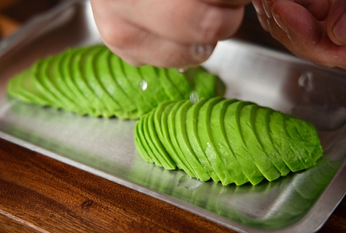 Fresh lemon juice being squeezed by hand over a tray of neatly arranged avocado slices.