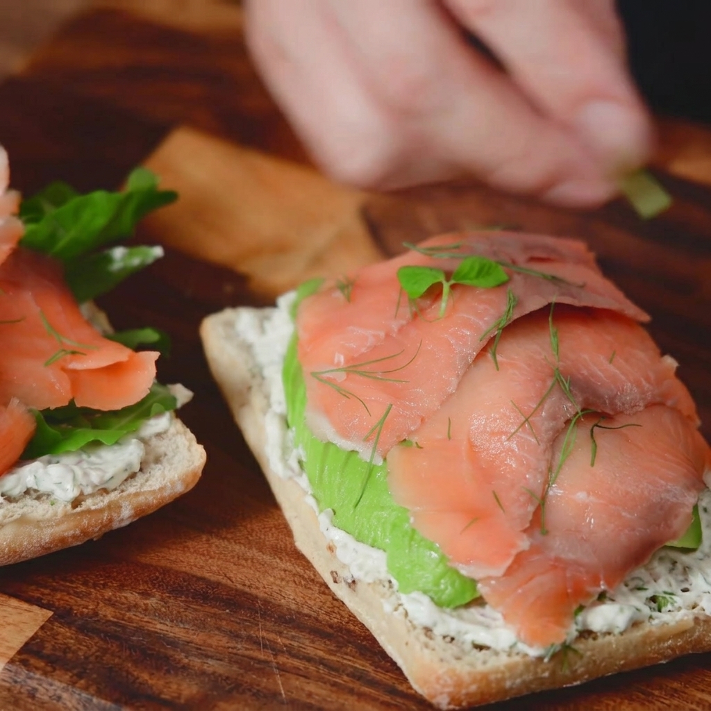 A macro photograph of fresh green dill sprigs and tiny microgreens being added as a garnish to the top of a smoked salmon sandwich.