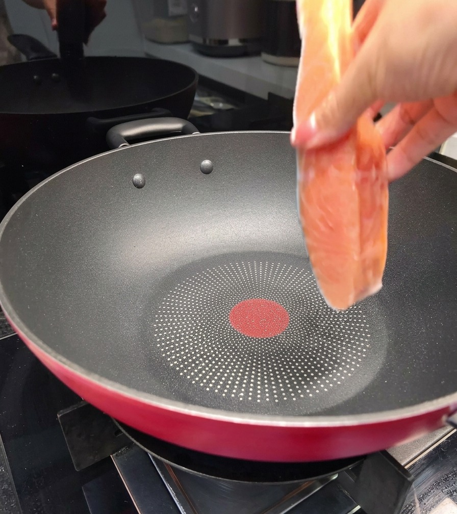 A hand placing a fresh salmon fillet into a large black non-stick pan on a stovetop.