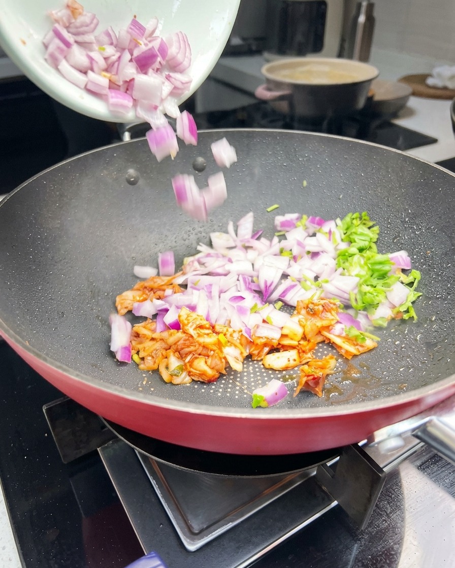 Chopped purple onions, green cilantro, and orange kimchi being tossed in a frying pan.