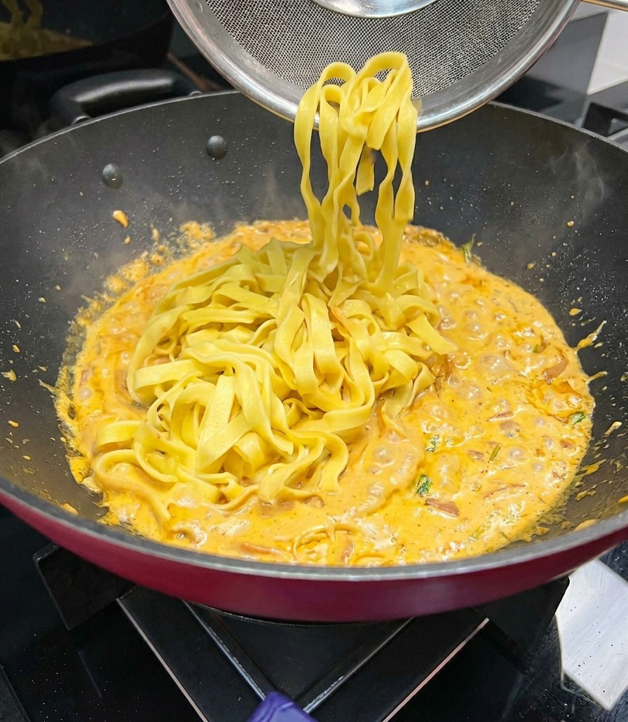 Wide fettuccine noodles being lifted with a mesh strainer and added to a pan of simmering orange cream sauce.