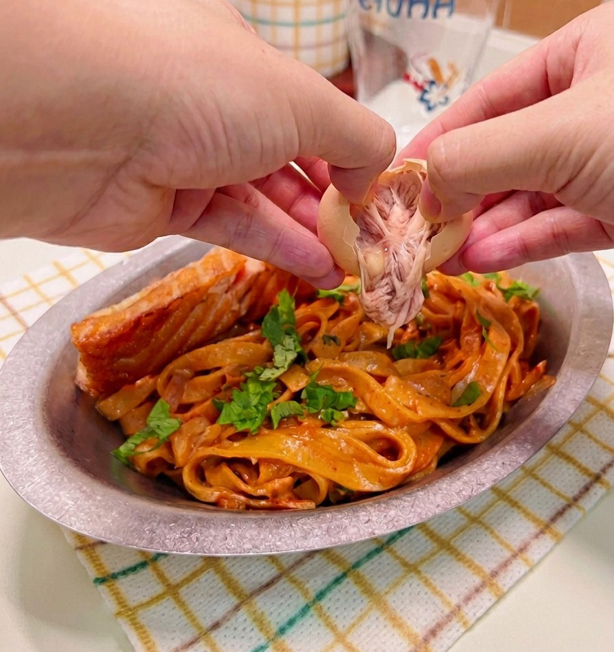 Hands carefully peeling and placing a soft-boiled egg over a plated dish of spicy cream pasta and seared salmon.