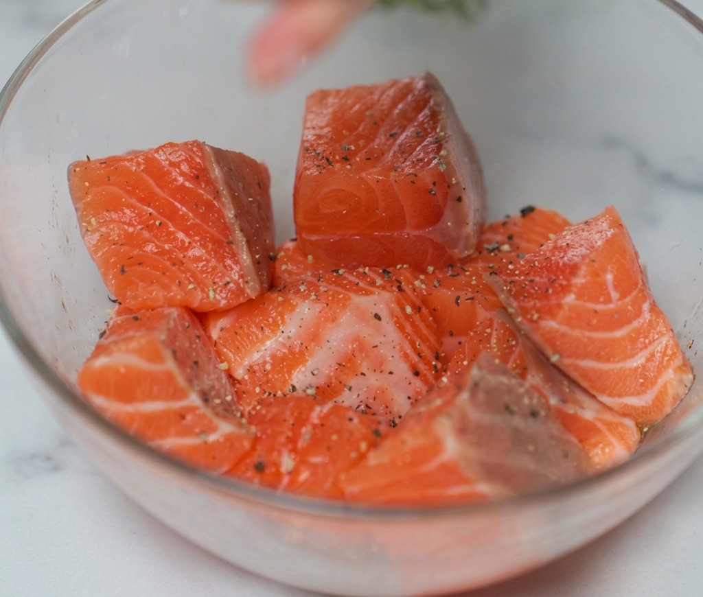 Raw salmon cubes in a glass bowl being sprinkled with salt and black pepper from a grinder.