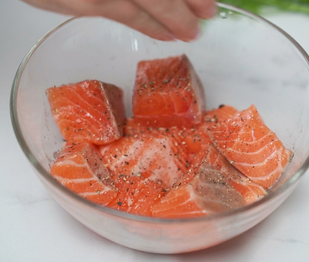 A hand squeezing fresh lemon juice onto seasoned salmon cubes in a clear bowl.