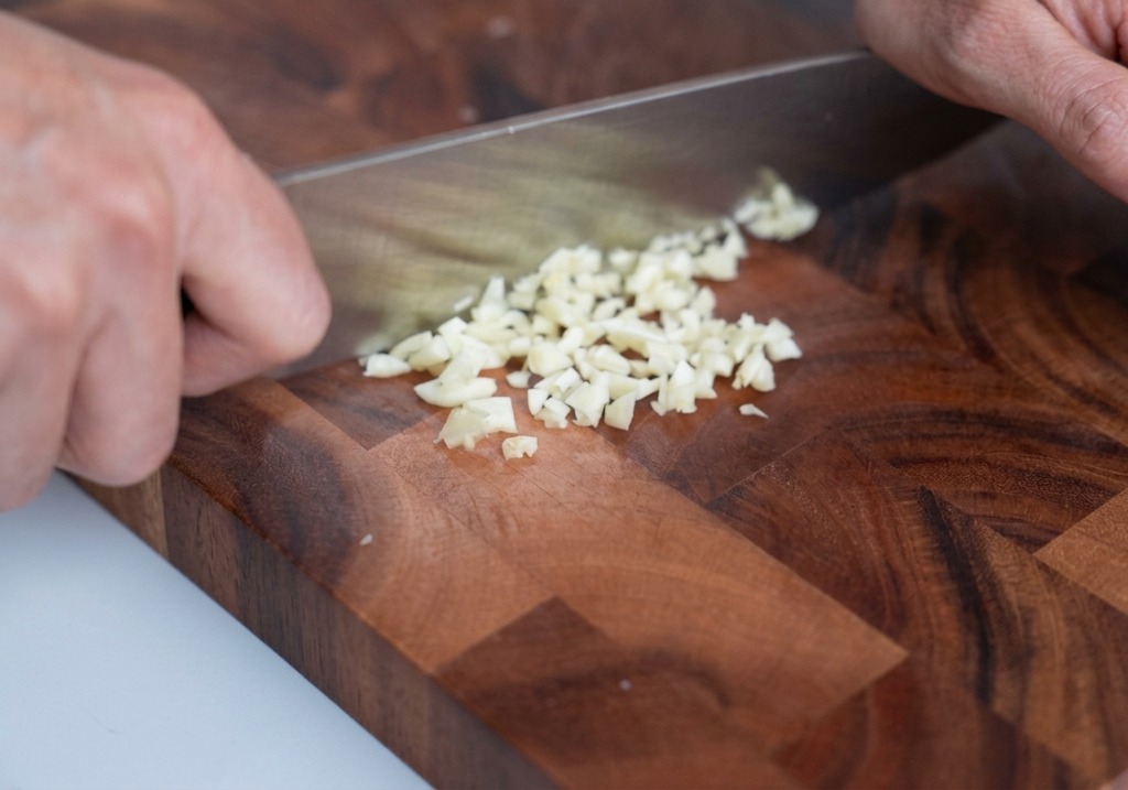 Finely minced garlic on a wooden cutting board with a chefs knife nearby.