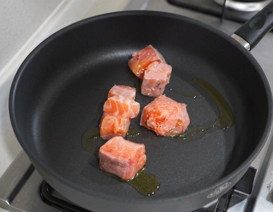 Cubed salmon pieces searing in a black non-stick pan with olive oil over a gas stove.