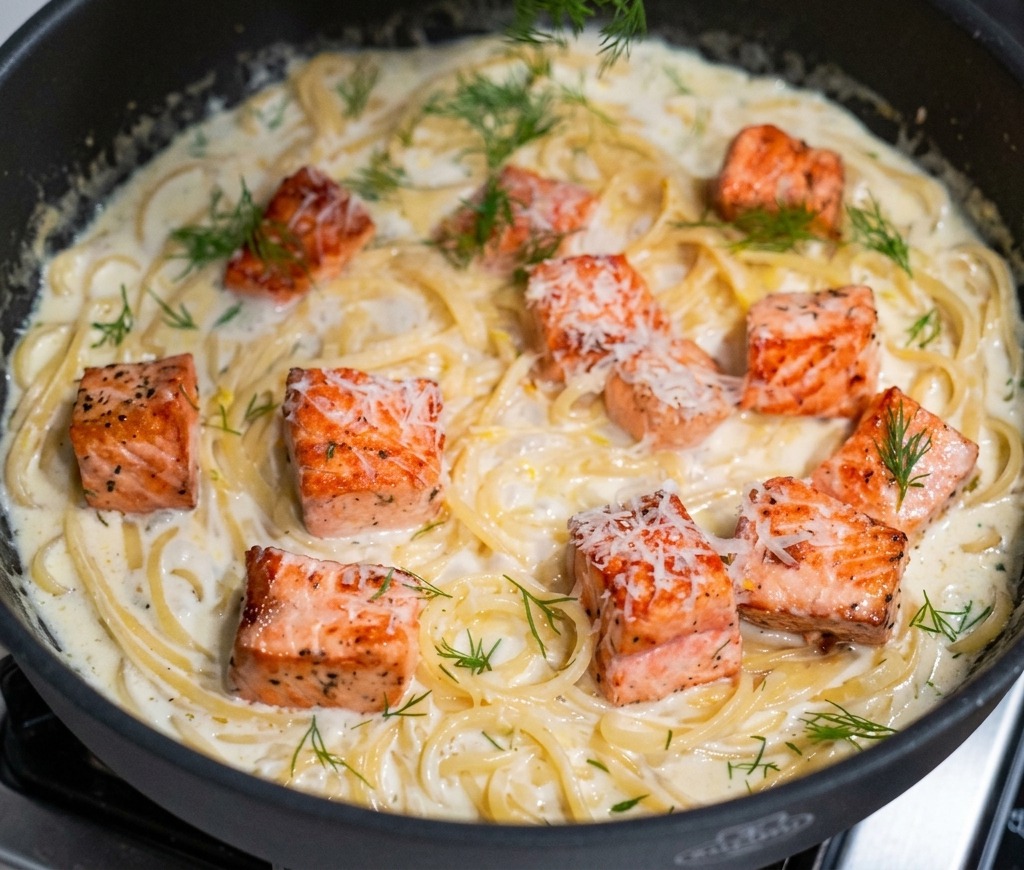 A hand sprinkling chopped green dill over a finished pan of creamy salmon pasta.