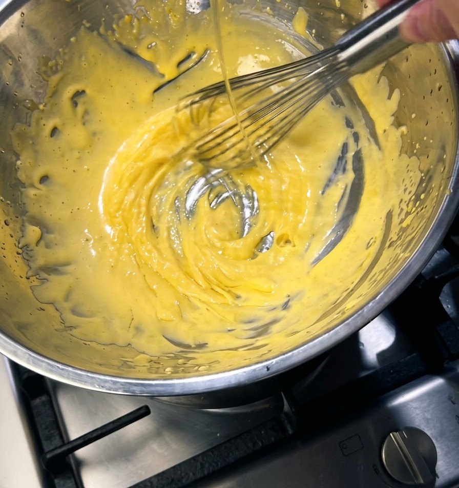 A metal whisk stirring a thick pale yellow mixture in a stainless steel bowl while a thin stream of golden butter is poured inside