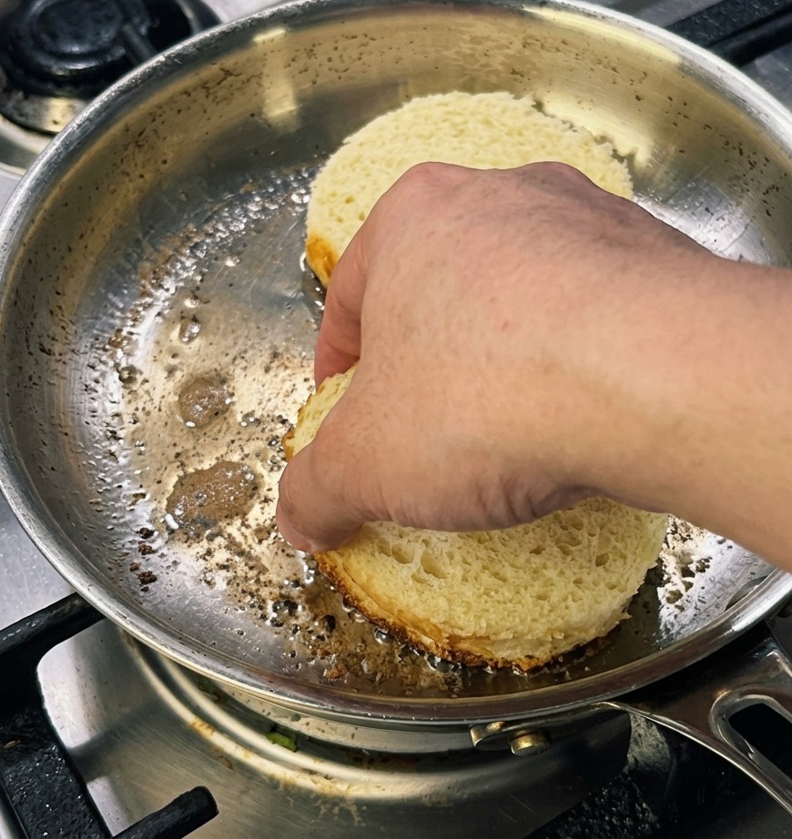A hand pressing down on two thick, round slices of brioche bread in a pan to achieve a golden-brown crust.