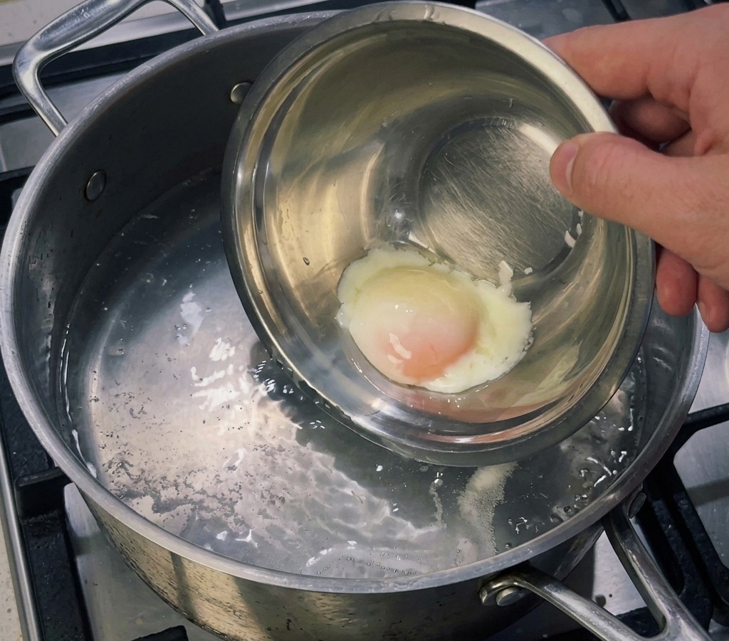 A small metal bowl containing a soft-cooked egg being lowered into a pot of simmering water to firm up the egg white.