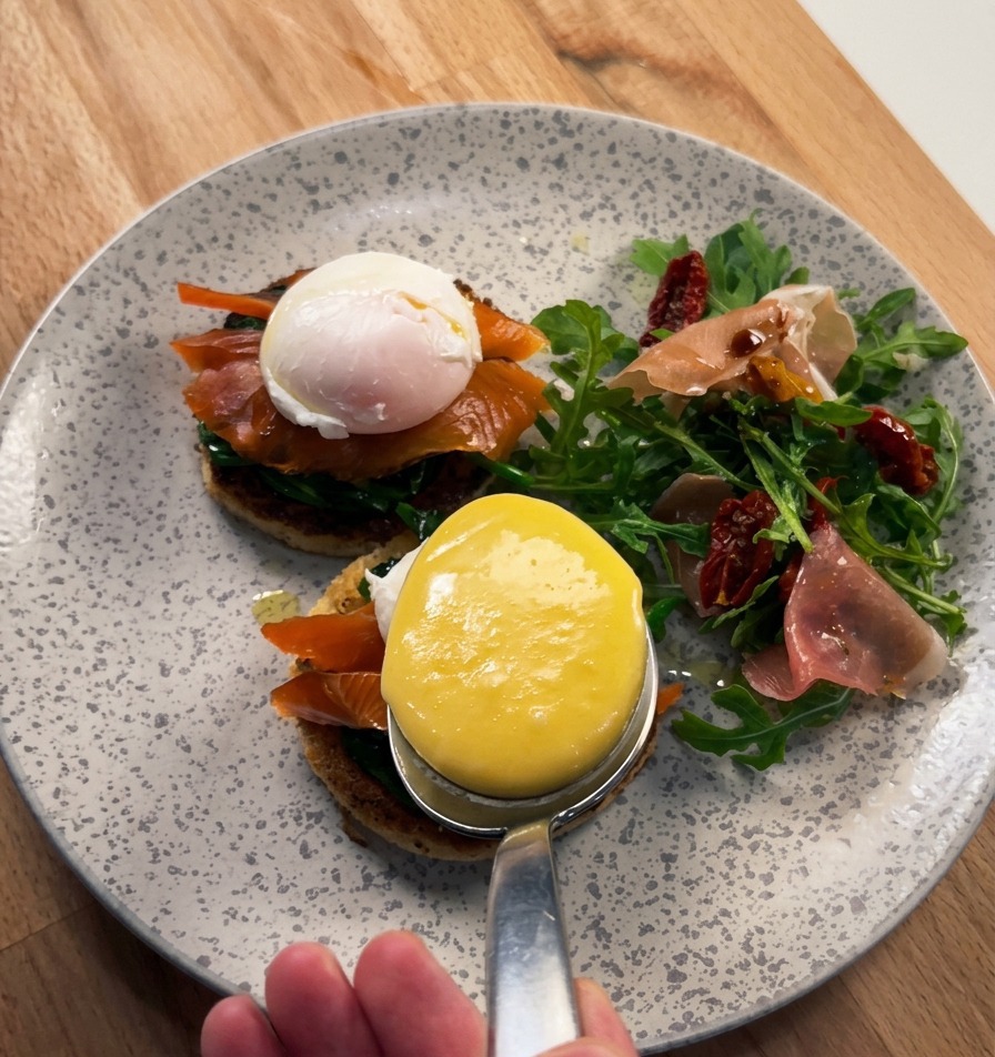 A close-up of a large spoon pouring thick, glossy yellow Hollandaise sauce over a poached egg resting on smoked salmon and brioche.