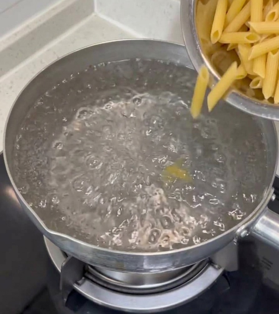 Dry penne pasta being poured from a stainless steel bowl into a pot of bubbling boiling water on a stovetop.
