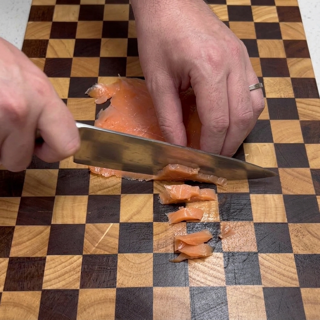 Chefs hands using a silver knife to cut flat pieces of smoked salmon into small squares on a brown and tan checkered cutting board.