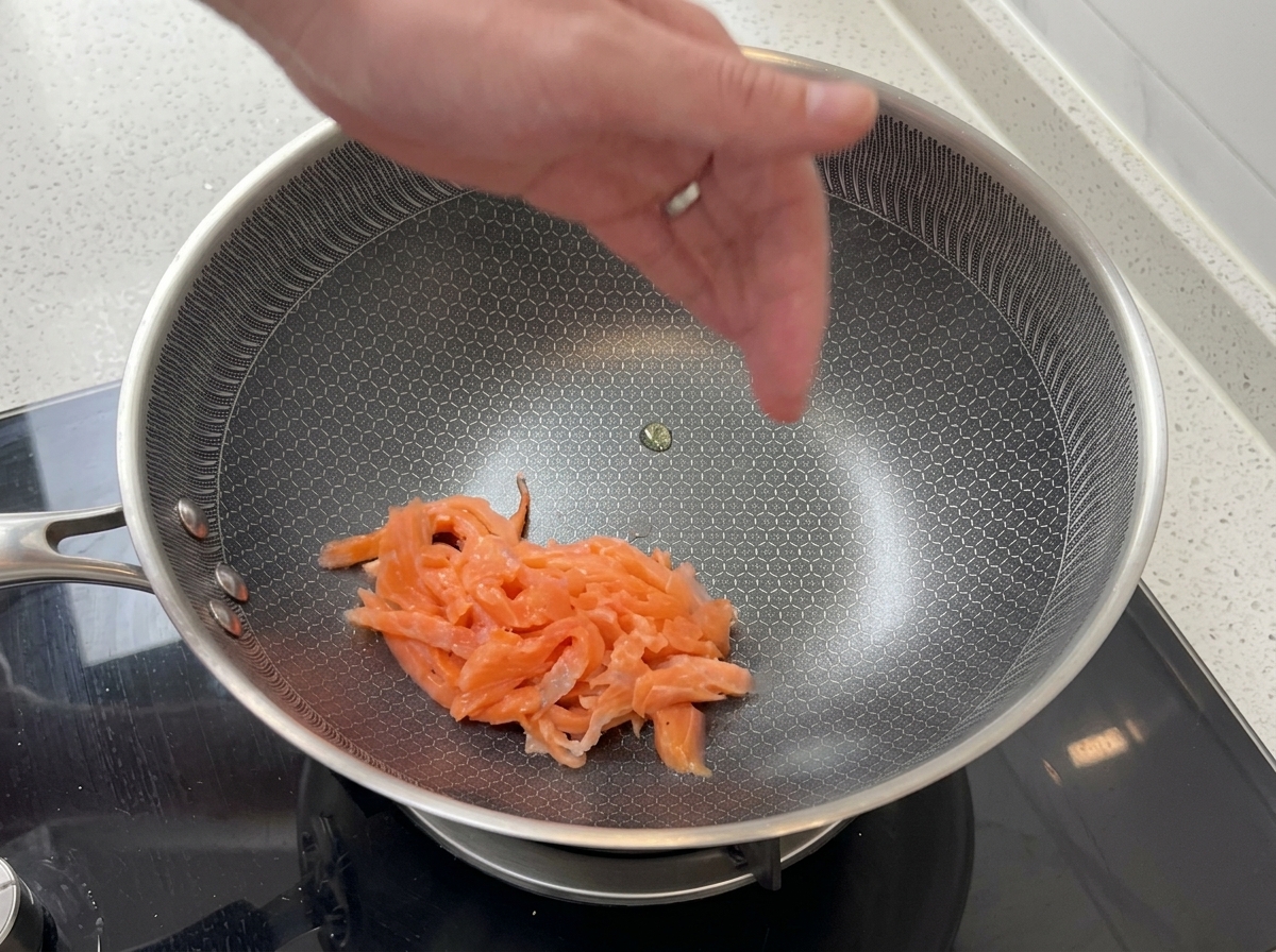 Slices of pink smoked salmon being tossed in a stainless steel frying pan with a textured hexagonal pattern.