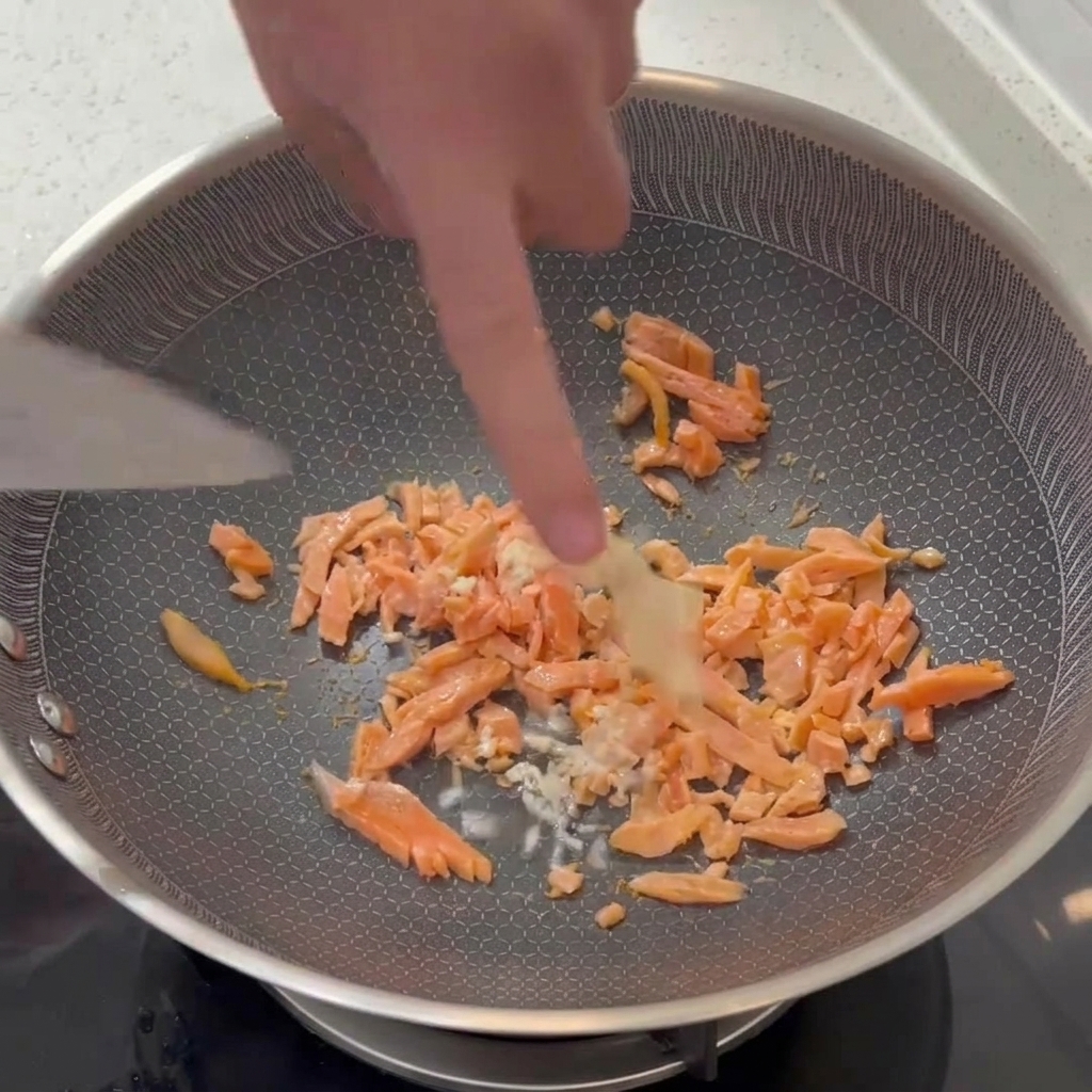 Minced white garlic being mixed into a pan of partially cooked smoked salmon pieces with a spatula.