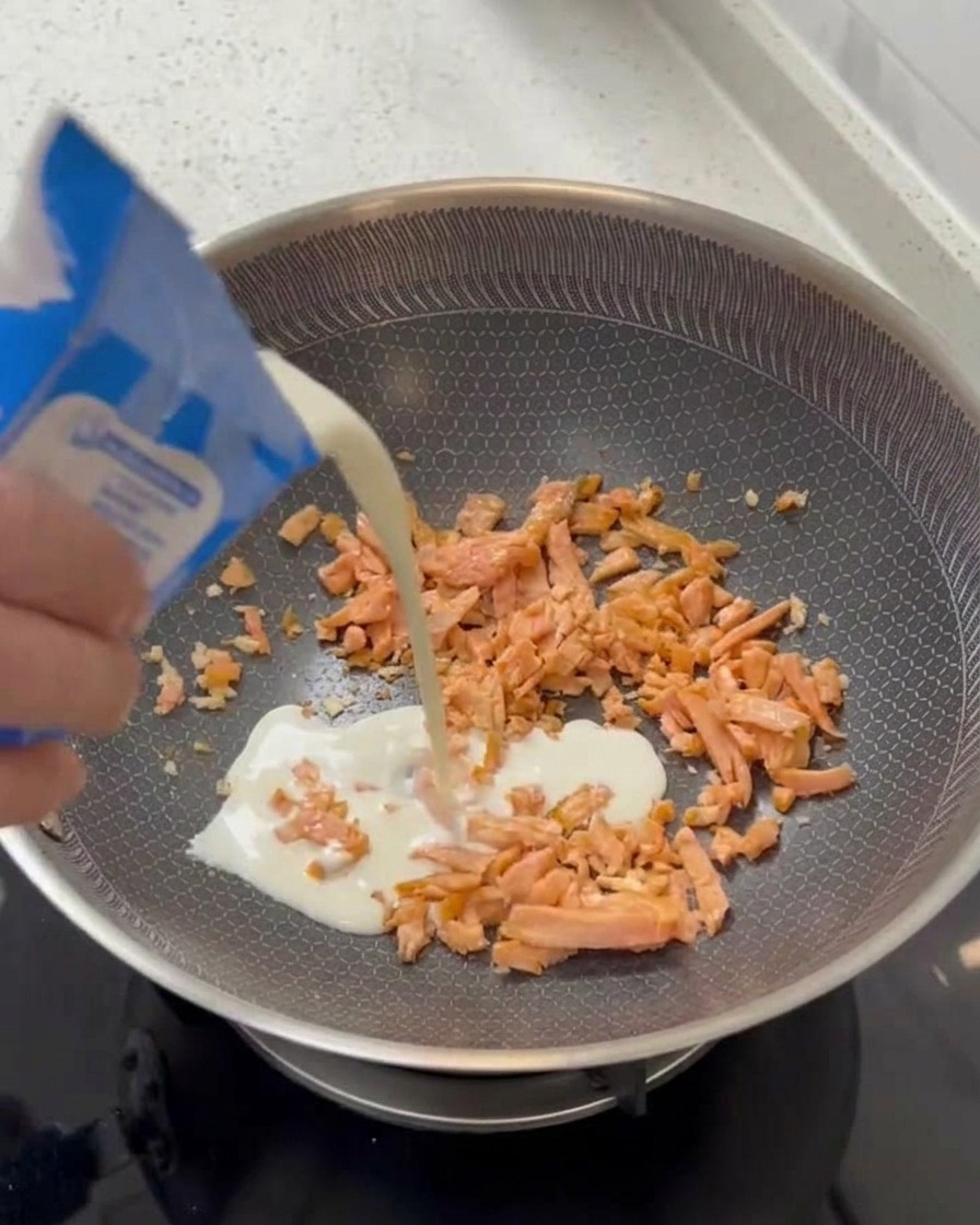 A hand pouring white light cream from a blue and white carton into a pan containing cooked salmon pieces.