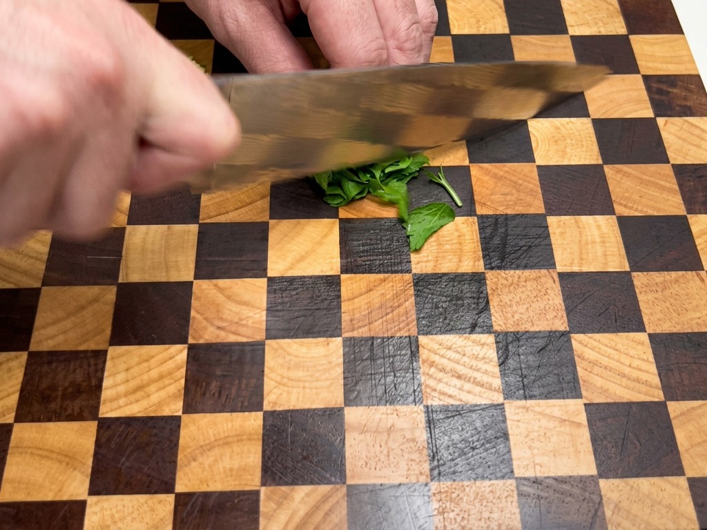 Close-up of hands using a chefs knife to finely chop fresh green parsley leaves on a checkered wooden cutting board.