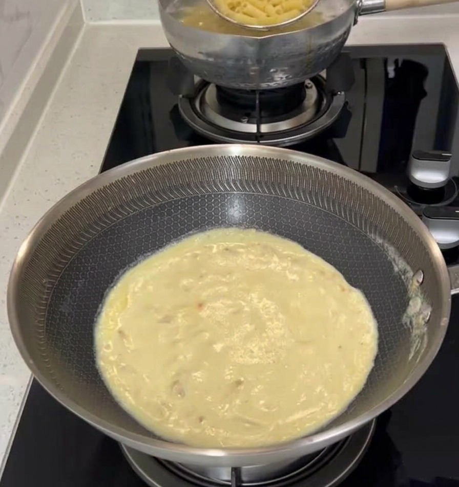 A metal spider strainer lifting cooked penne pasta from a boiling pot and transferring it into a pan filled with creamy white sauce on a stovetop.