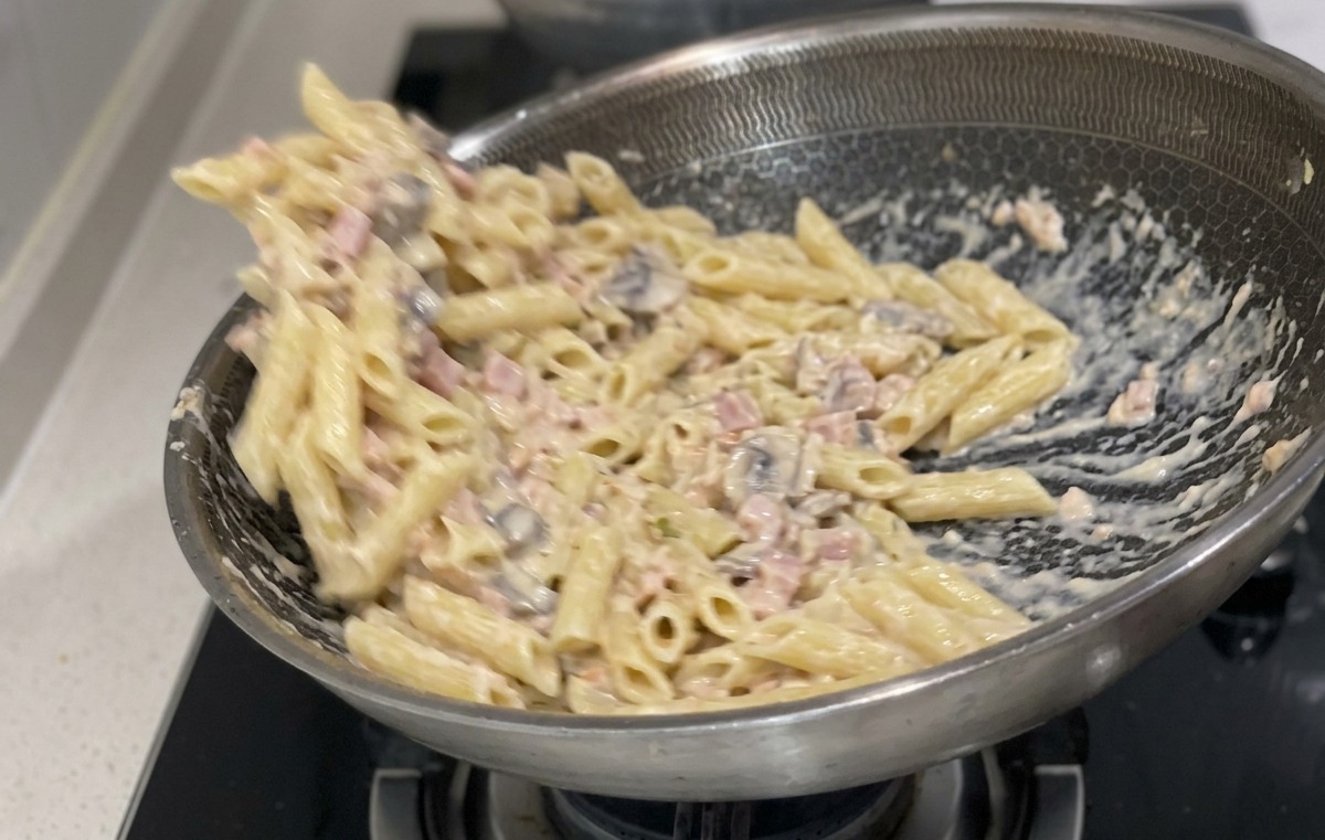 Cooked penne pasta being tossed and mixed with a creamy white sauce and salmon pieces in a large stainless steel frying pan.
