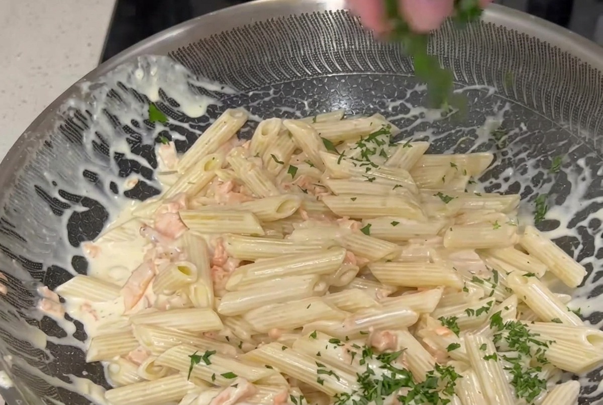 A hand sprinkling finely chopped green parsley over a large pan of penne pasta tossed in a creamy white sauce with pink smoked salmon pieces.