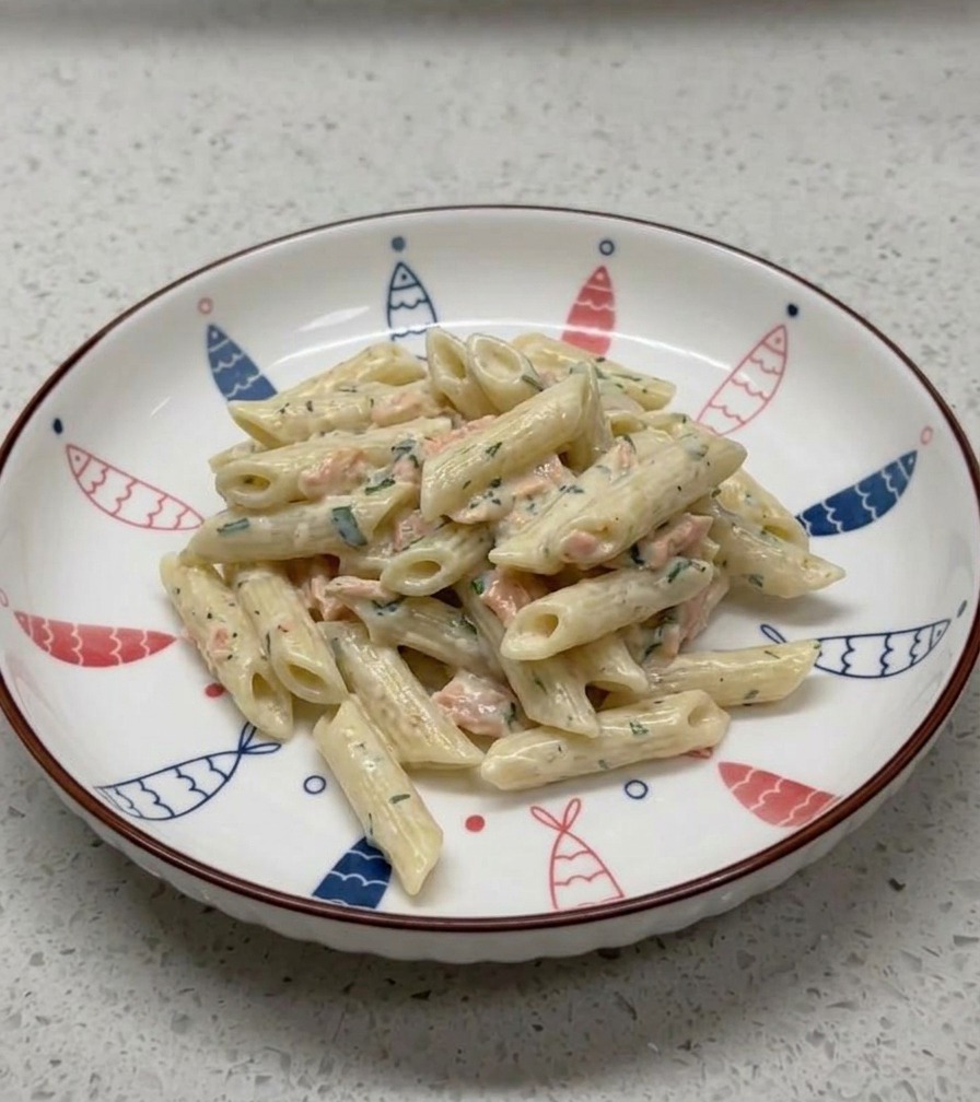 A serving of creamy salmon penne pasta plated neatly in a white ceramic bowl decorated with a blue and red fish pattern around the rim.