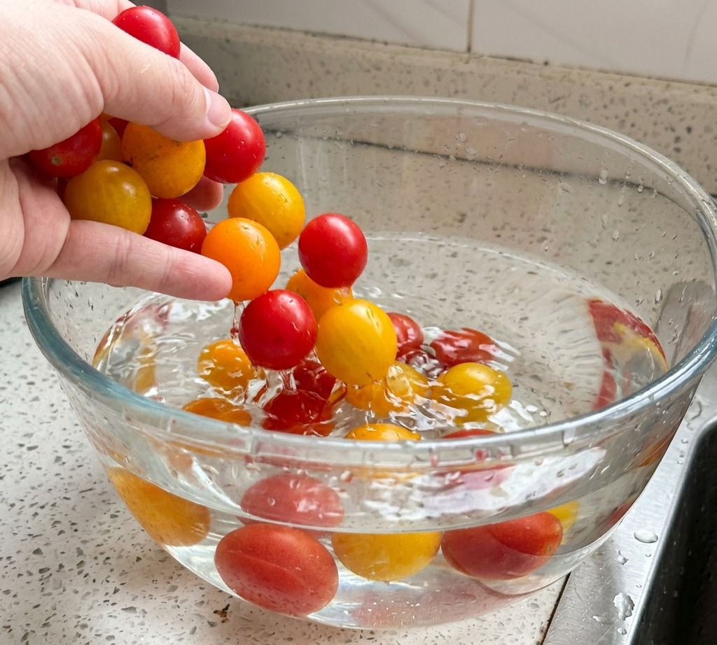 A persons hand washing a mix of vibrant red and yellow cherry tomatoes in a large glass bowl filled with water.